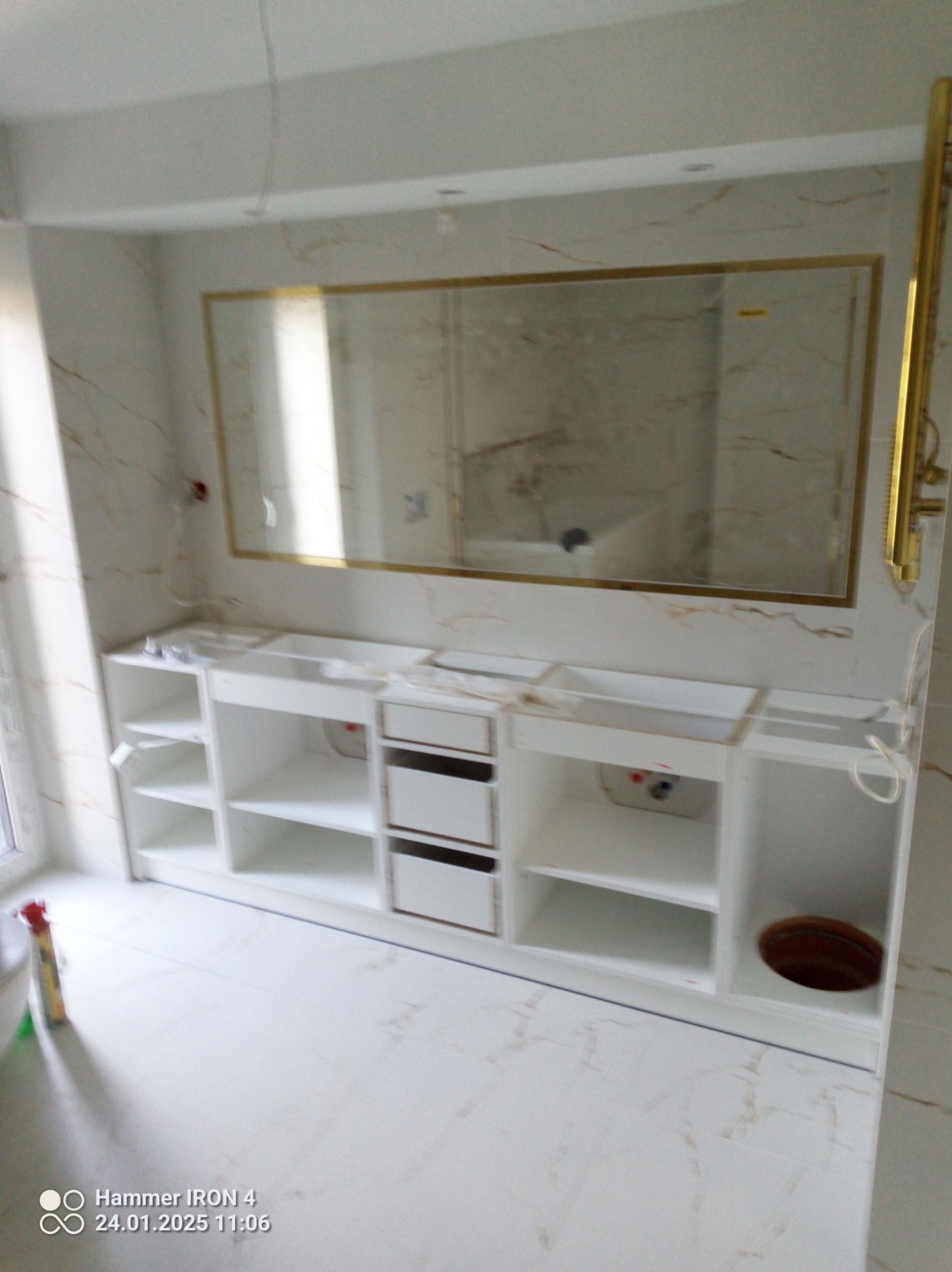 Biała szafka pod umywalkę z szufladami i półkami w łazience wykończonej marmurem, nad nią lustro w złotej ramie. Widoczne elementy instalacji.