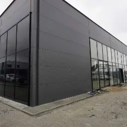 Nowoczesny budynek biurowy z szarymi panelami elewacyjnymi i dużymi, panoramicznymi oknami z czarnymi ramami, widok z zewnątrz na parking i ulicę.