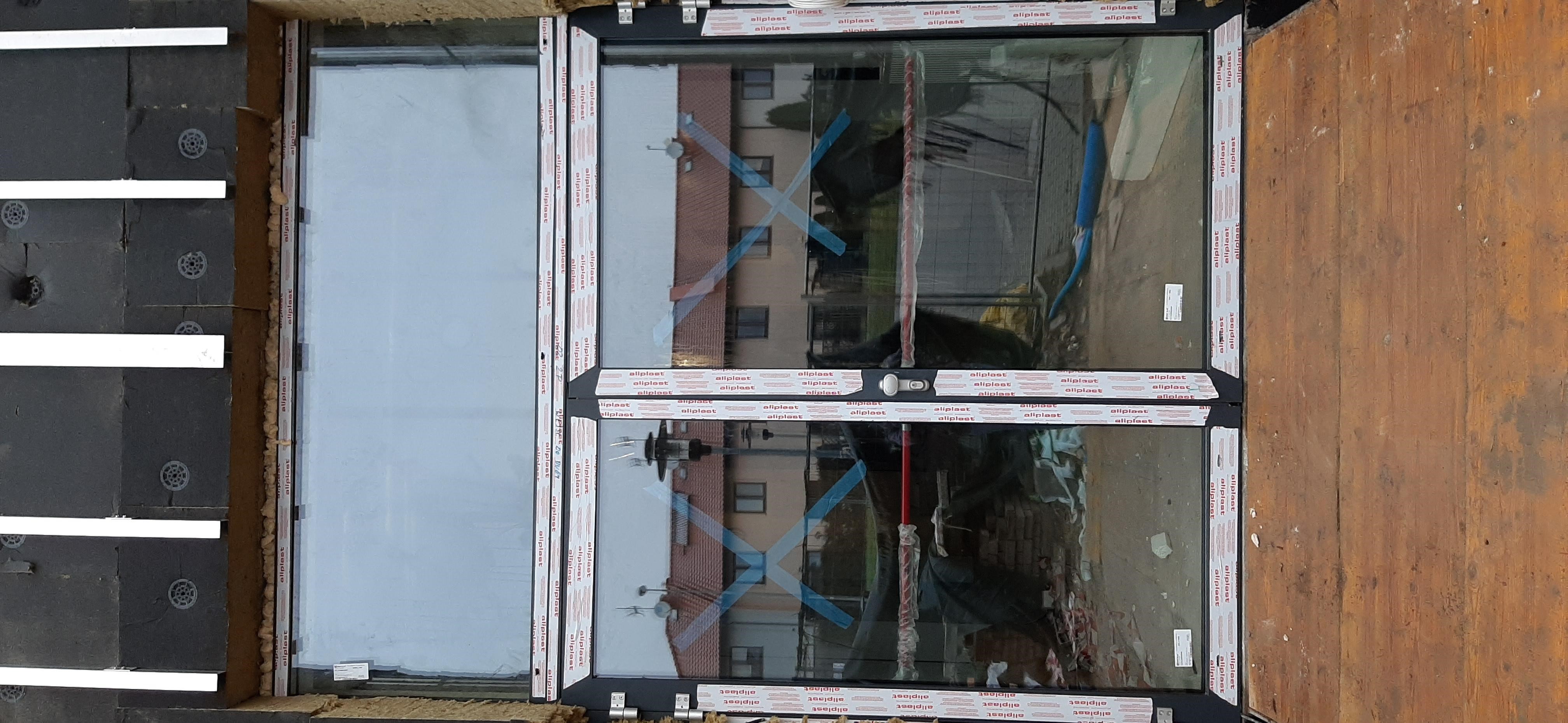 Nowe okno PCV z folią ochronną zamontowane w surowym otworze okiennym, widoczne materiały izolacyjne i odblask budynku w szybie.