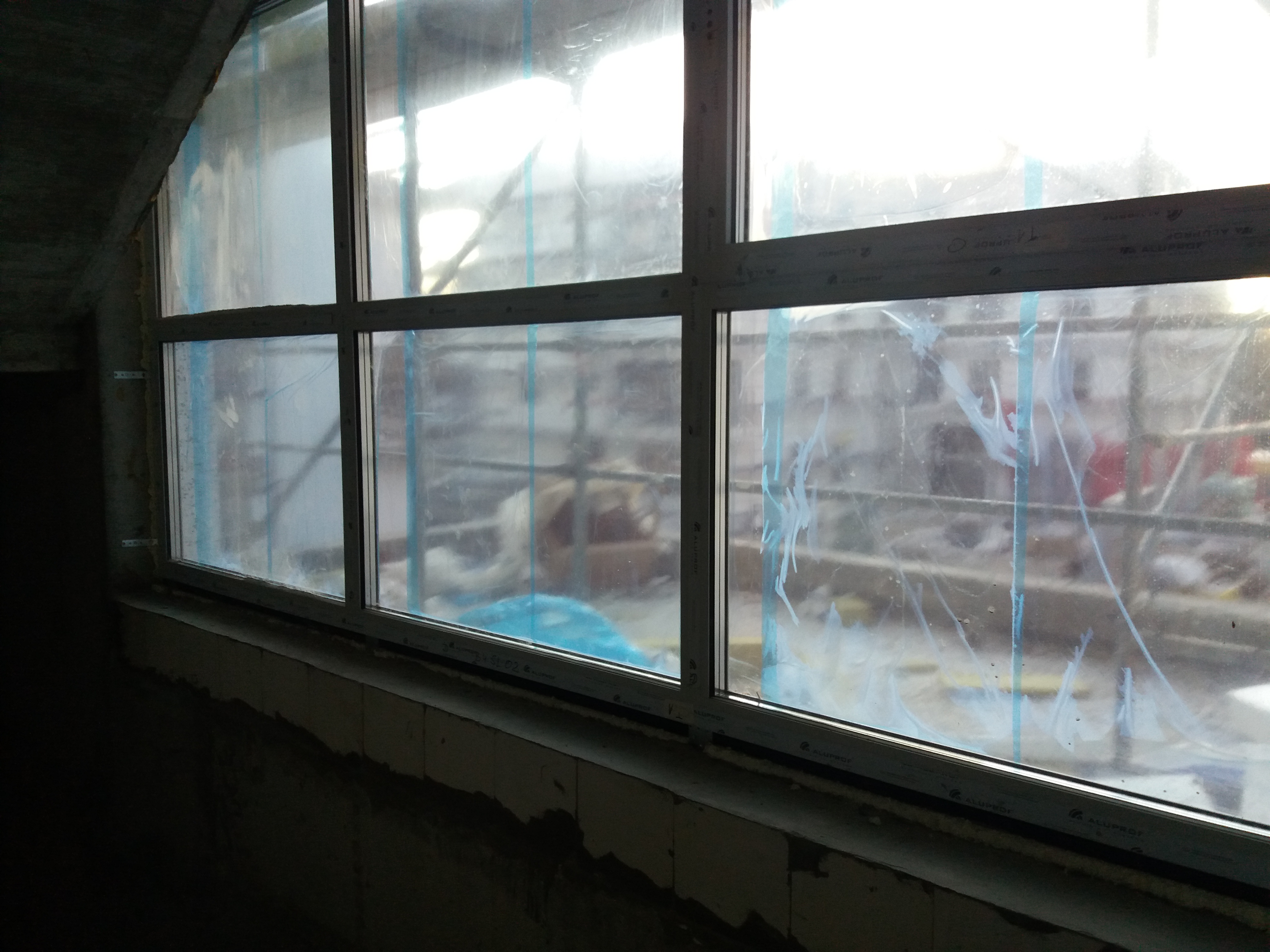 Zamontowane okno PCV, zabezpieczone folią ochronną, widoczne ślady po kleju, surowy stan wykończenia wnętrza.