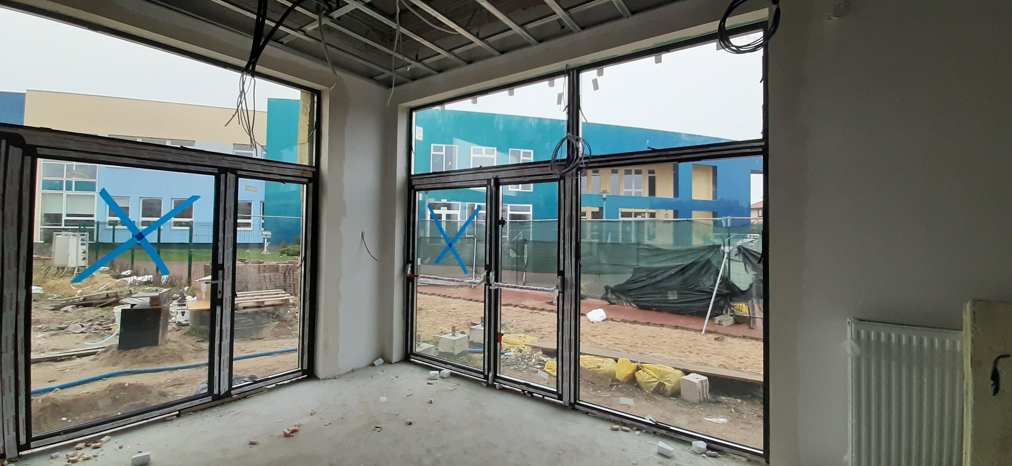 Wnętrze w trakcie wykańczania z zamontowanymi oknami aluminiowymi z widocznym niebieskim krzyżem na szybie zabezpieczającym przed uszkodzeniem, na zewnątrz widoczny budynek w kolorach niebieskim...