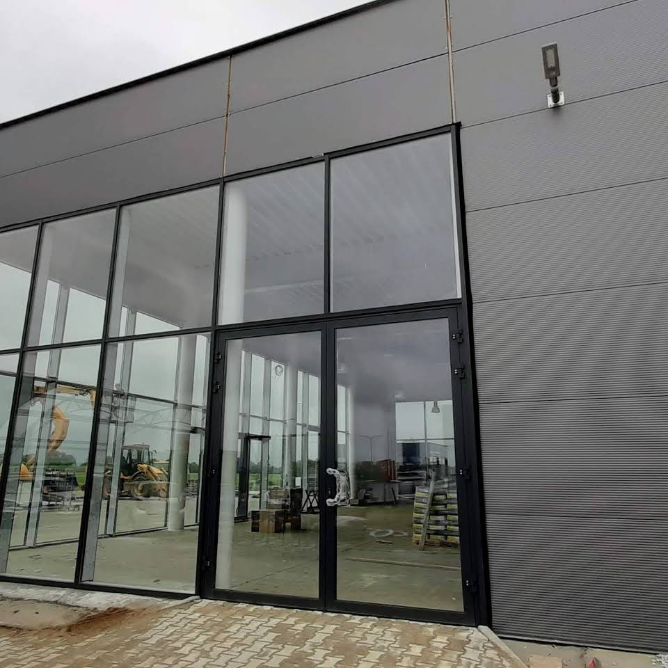 Nowoczesne, przeszklone wejście do budynku przemysłowego z czarnymi ramami, widoczne maszyny budowlane wewnątrz, szara fasada z blachy trapezowej.
