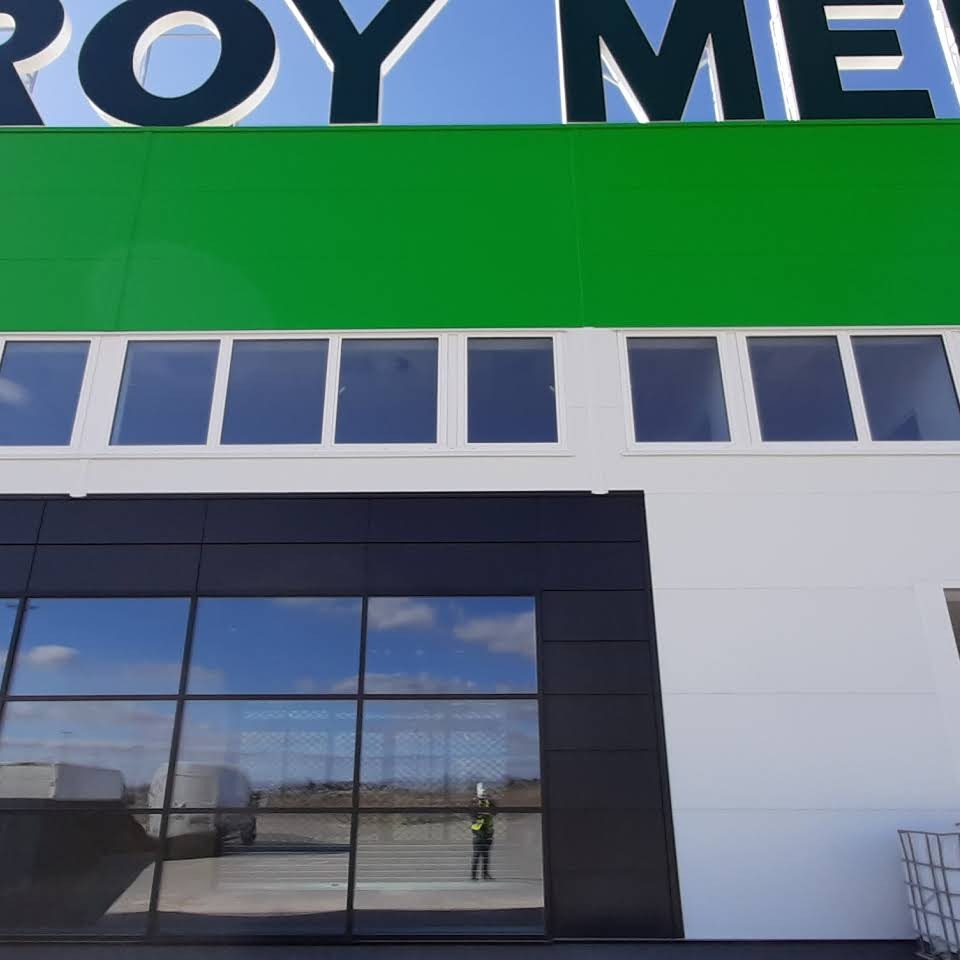 Fasada budynku z nowoczesnymi oknami aluminiowymi; w dolnych oknach odbija się niebo i pracownik w kamizelce odblaskowej.