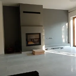 Nowoczesny salon z szarymi ścianami, kominkiem z czarną obudową i jasnymi, kwadratowymi płytkami podłogowymi, częściowo posprzątany odkurzaczem.