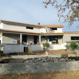 Dwukondygnacyjny dom murowany w trakcie budowy, z częściowo położonym dachem z czerwonej dachówki. Dwóch robotników pracuje na dachu, a w tle widoczne drzewo z uschniętymi liśćmi i błękitne niebo.