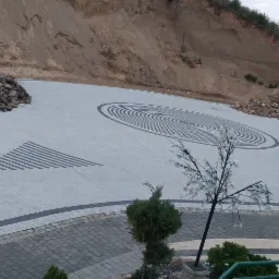 Rozległy teren z kostki brukowej z geometrycznymi wzorami: okrągłe koncentryczne linie i trójkąt z pasków, na tle skarpy ziemnej i sztucznego drzewka z białymi kwiatami.