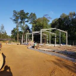 Stalowa konstrukcja budynku w trakcie montażu, widoczna na tle zieleni drzew i błękitnego nieba; na pierwszym planie przygotowany teren z piaskiem i sprzęt budowlany.