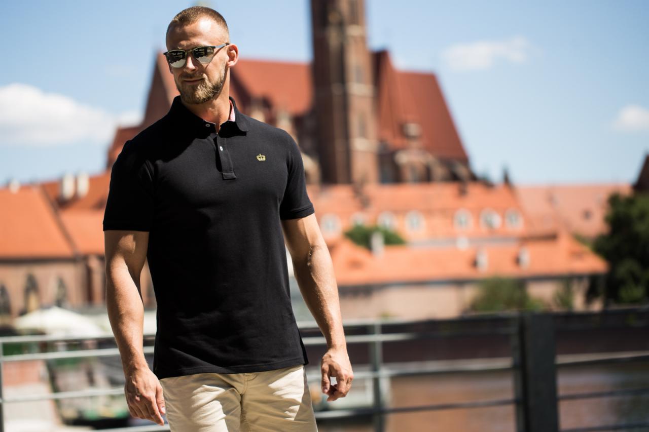 Mężczyzna w czarnej koszulce polo z koroną, okularach przeciwsłonecznych, na tle ceglanych budynków i niebieskiego nieba.