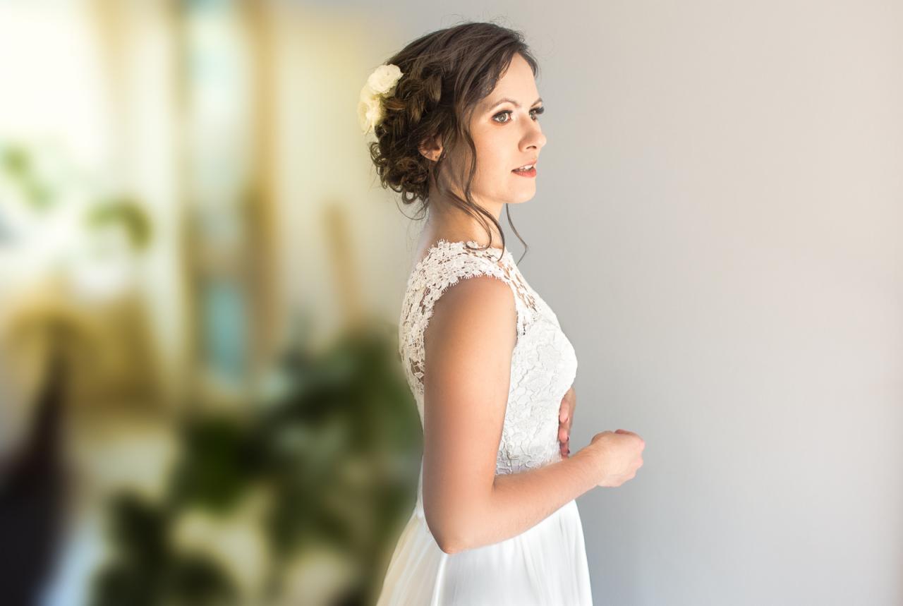 Panna młoda w białej koronkowej sukni, profil, z kwiatami we włosach, zamyślona, delikatny makijaż.