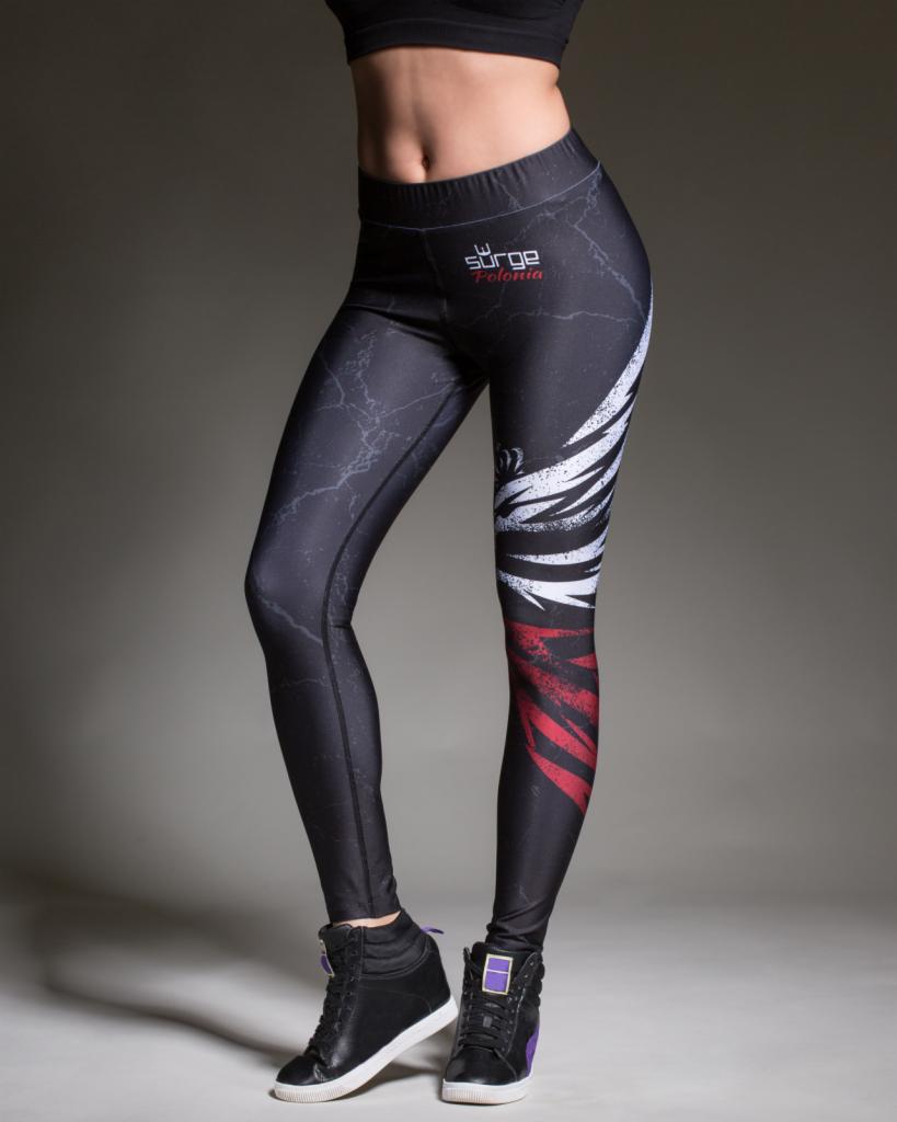 Kobieta w czarnych legginsach z nadrukiem błyskawic i skrzydeł, pozująca w czarnych butach sportowych na koturnie