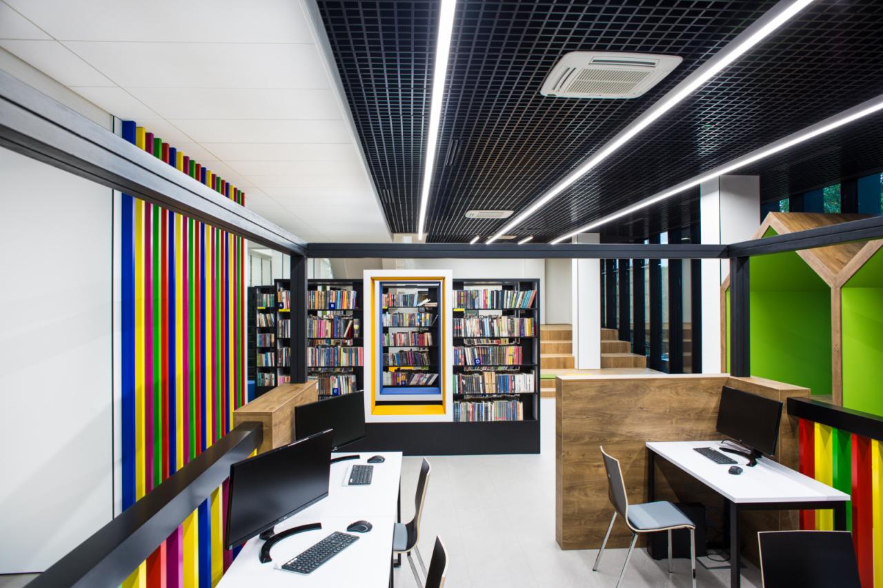 Wnętrze nowoczesnej biblioteki z regałami pełnymi książek, stanowiskami komputerowymi i elementami dekoracyjnymi w postaci kolorowych pionowych pasków.