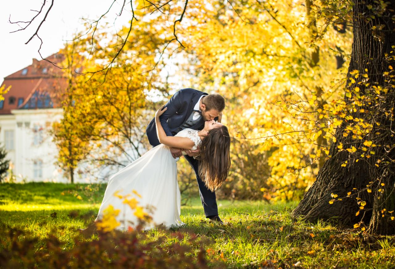 Pocałunek młodej pary w jesiennej scenerii parku we Wrocławiu, panna młoda w białej sukni, pan młody w granatowym garniturze, w tle klasycystyczny budynek.