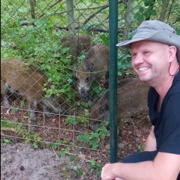 Wintech - Uśmiechnięty mężczyzna w kapeluszu kuca przed ogrodzeniem, za którym widać dziki w lesie.