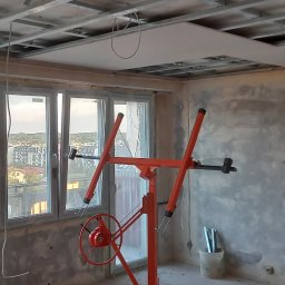 Wintech - Pomarańczowy podnośnik do płyt karton gips, ustawiony w surowym wnętrzu z oknem, widoczny montaż płyty na metalowym stelażu sufitowym.