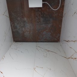 Wintech - Wykończona kabina prysznicowa z odpływem liniowym, ściany wyłożone płytkami imitującymi marmur i rdzawy kamień, widoczna słuchawka prysznicowa.