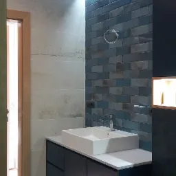 Nowoczesna łazienka z umywalką nablatową na szafce w kolorze granatowym, podświetloną ścianą z niebieskich płytek cegiełek i szarą podłogą z płytek wielkoformatowych.