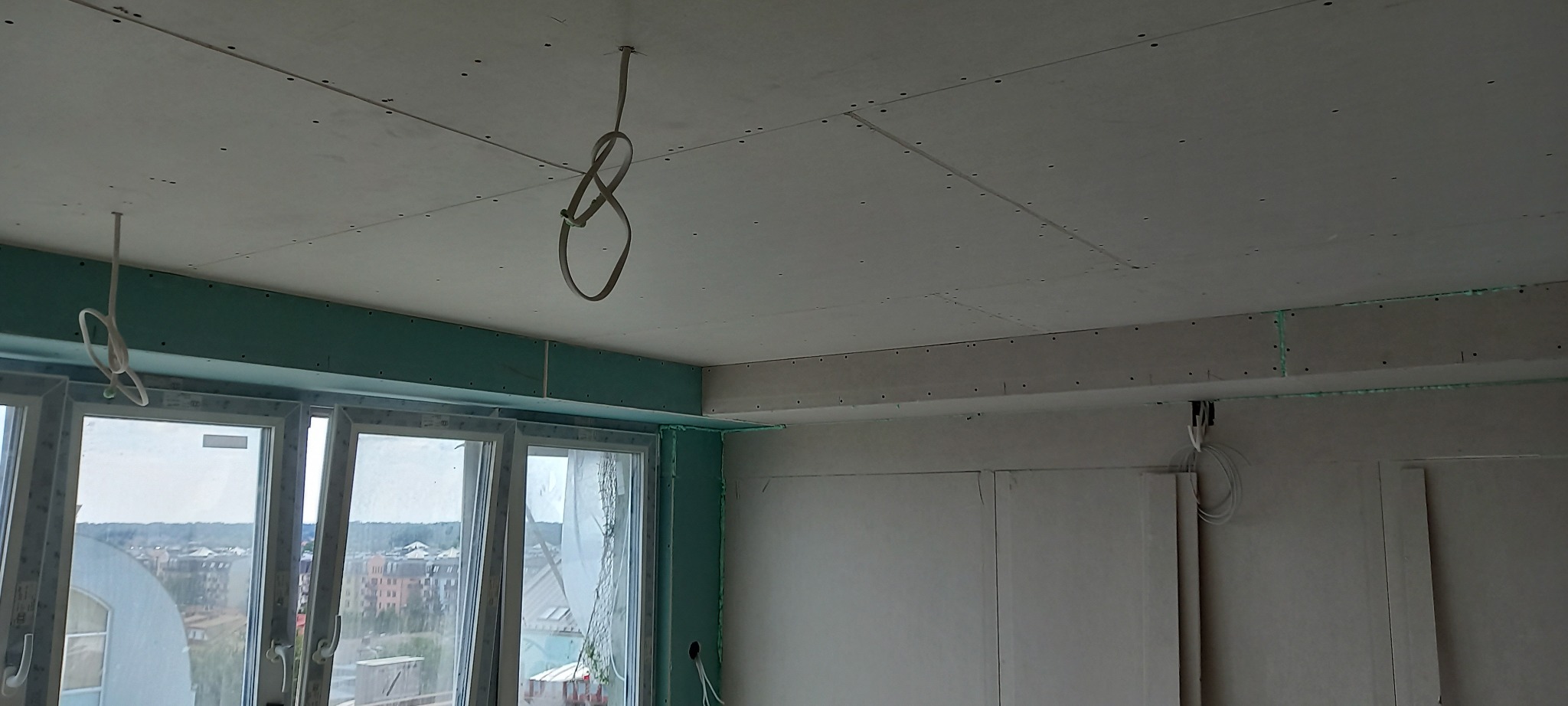 Montaż płyt kartonowo-gipsowych na suficie i ścianach w nowym mieszkaniu, widoczne kable elektryczne wystające z sufitu i zielone płyty GK przy oknach.