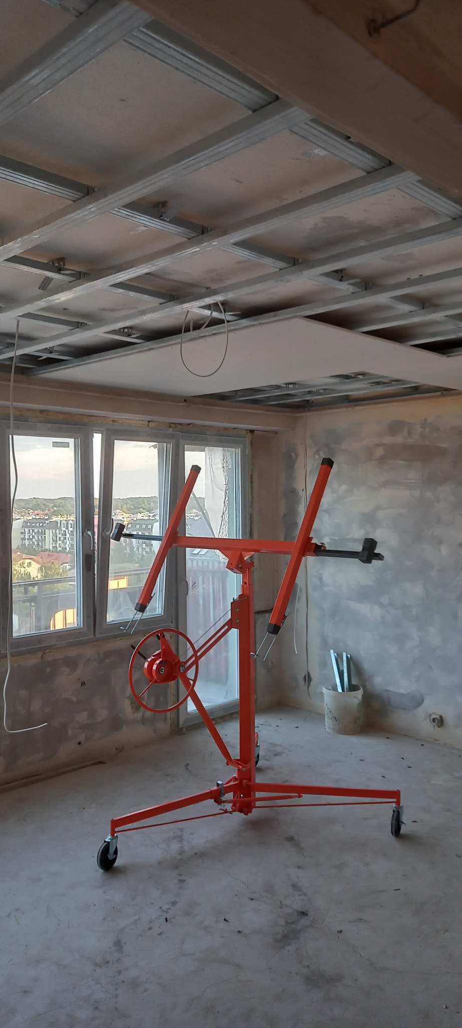 Pomarańczowy podnośnik do płyt karton gips, ustawiony w surowym wnętrzu z oknem, widoczny montaż płyty na metalowym stelażu sufitowym.