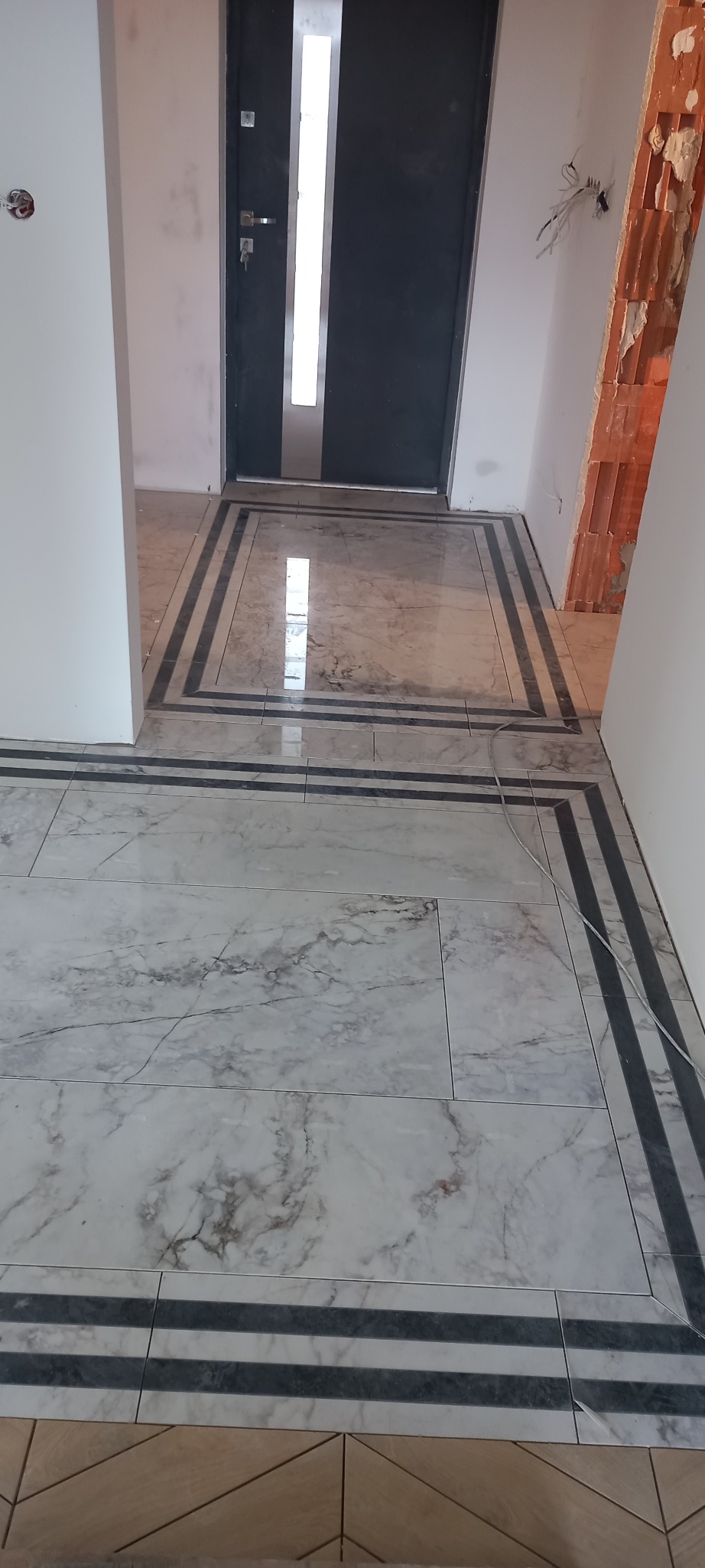 Ułożona marmurowa podłoga z dekoracyjnymi czarnymi paskami w korytarzu, widoczne niedokończone ściany i instalacja elektryczna.