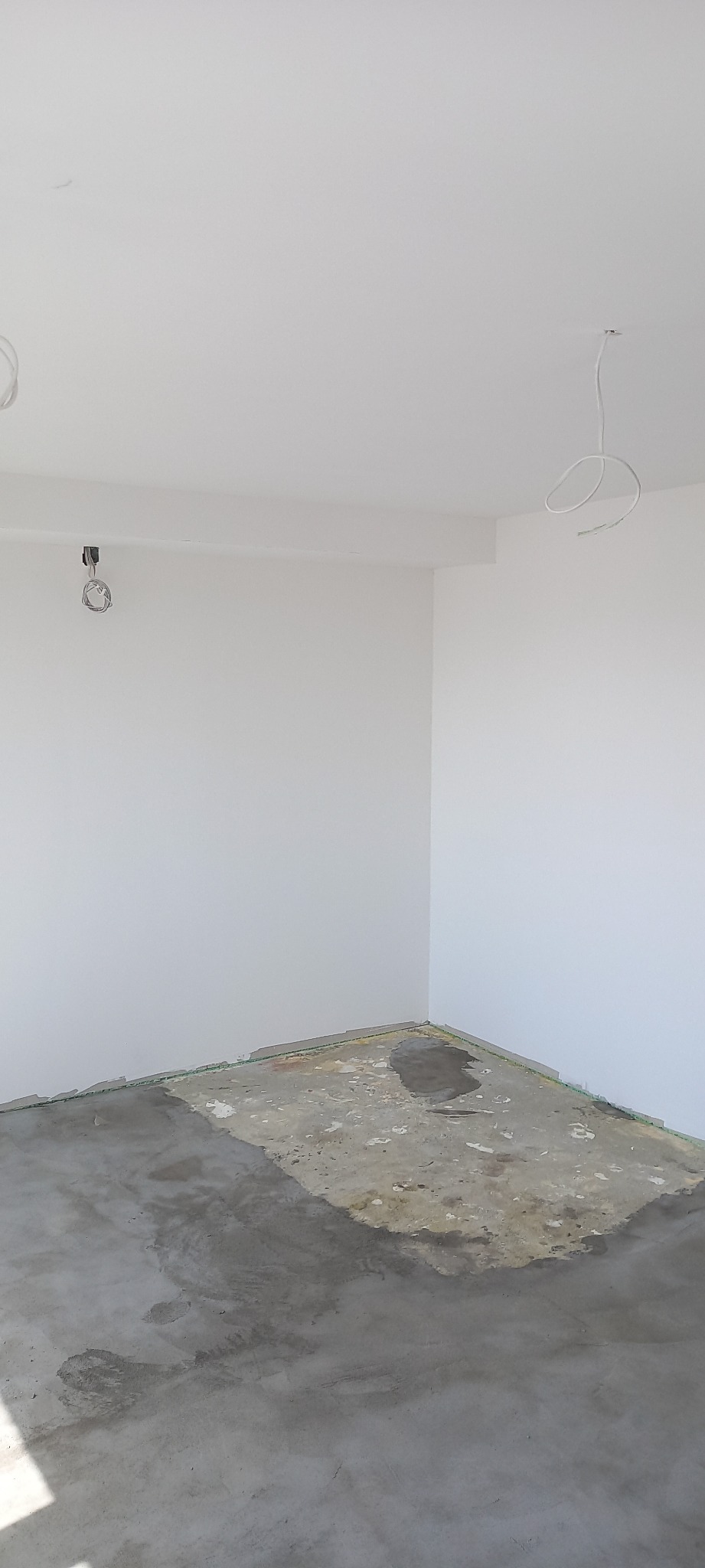 Surowy betonowy podkład podłogowy w pustym, jasnym pokoju z białymi ścianami, przygotowany do dalszych prac wykończeniowych, widoczne wystające przewody elektryczne.