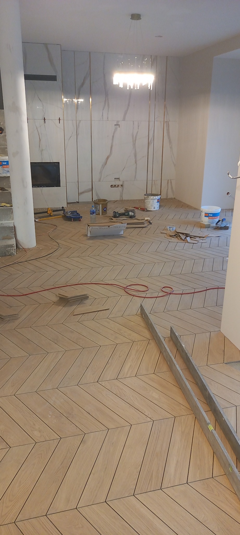 Układanie parkietu ceramicznego w jodełkę francuską w jasnym pomieszczeniu z marmurowymi ścianami, widoczne narzędzia glazurnicze i poziomice.