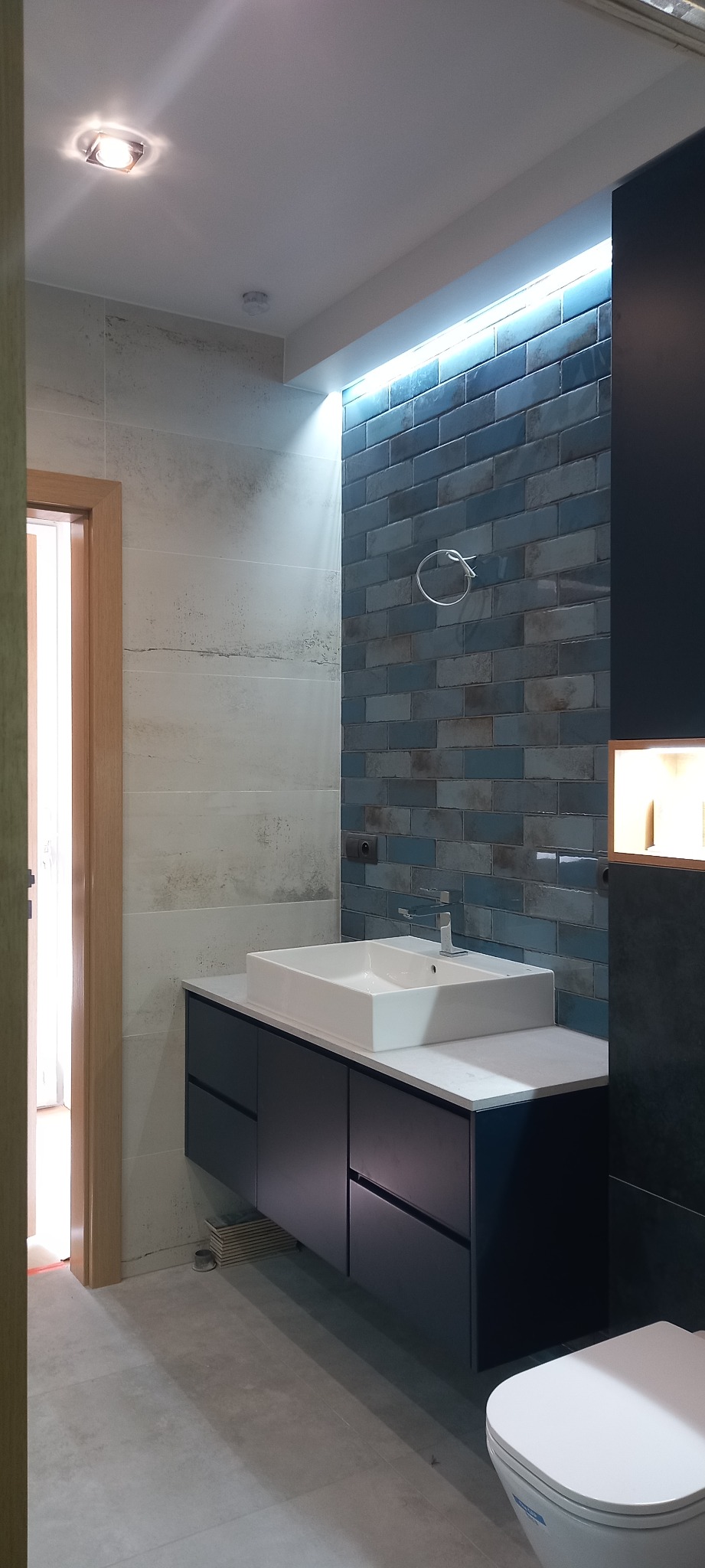 Nowoczesna łazienka z umywalką nablatową na szafce w kolorze granatowym, podświetloną ścianą z niebieskich płytek cegiełek i szarą podłogą z płytek wielkoformatowych.
