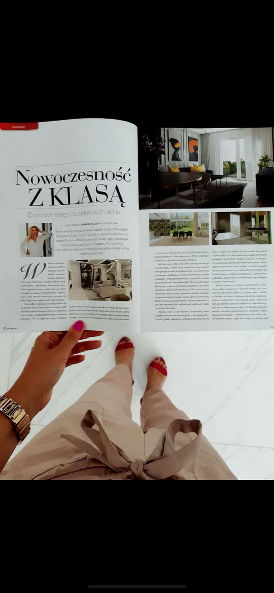 Otwarta gazeta z artykułem o aranżacji wnętrz, prezentująca zdjęcia nowoczesnych domów. Widoczne nogi osoby trzymającej gazetę, marmurowa podłoga.