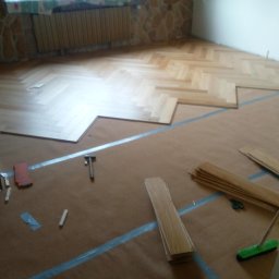 Mariusz Kujawski M-BUD - Układanie parkietu w jodełkę na brązowym podkładzie. Widoczne narzędzia: młotek, miarka, nożyczki. Część podłogi już wykończona, reszta w trakcie prac.