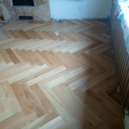 Mariusz Kujawski M-BUD - Ułożona podłoga z paneli w jodełkę, widoczne narzędzia i zabezpieczony grzejnik. Częściowo ułożona podłoga w pokoju z kominkiem.