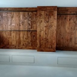 Mariusz Kujawski M-BUD - Drewniana ściana z przesuwnymi drzwiami w stylu rustykalnym, uzupełniona metalowymi rurami. Biała cegła i zabudowa z szufladami w jasnym wnętrzu.