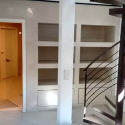 Mariusz Kujawski M-BUD - Wykończone wnętrze z spiralnymi schodami, wnękami w ścianie i otwartymi drzwiami. Minimalistyczny design, jasne kolory, ceglana ściana w tle.