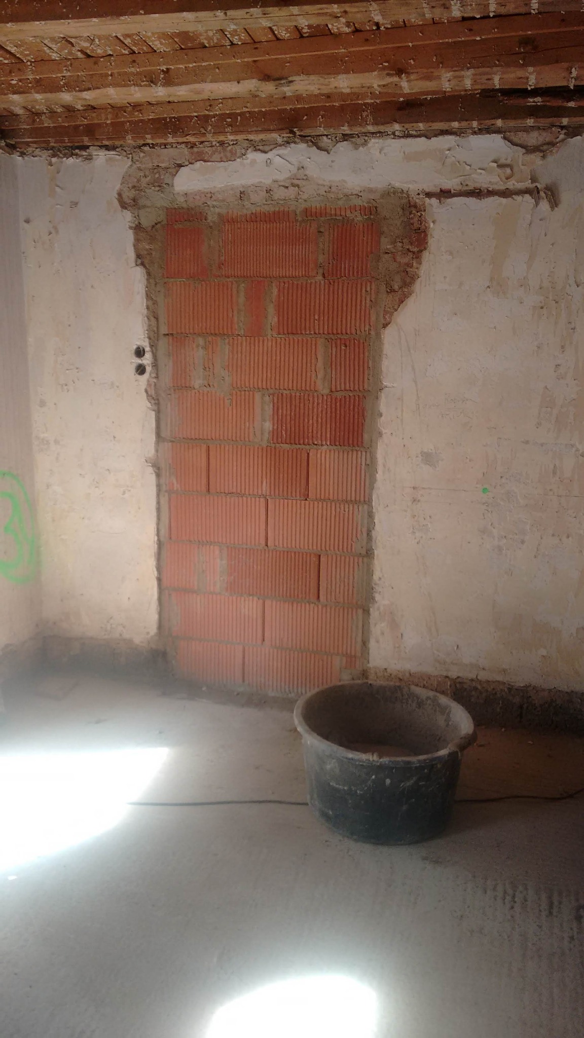 Murowane otwarcie w ścianie, widoczne ślady po kuciu, surowy stan, wiadro na podłodze, przygotowanie do dalszych prac.