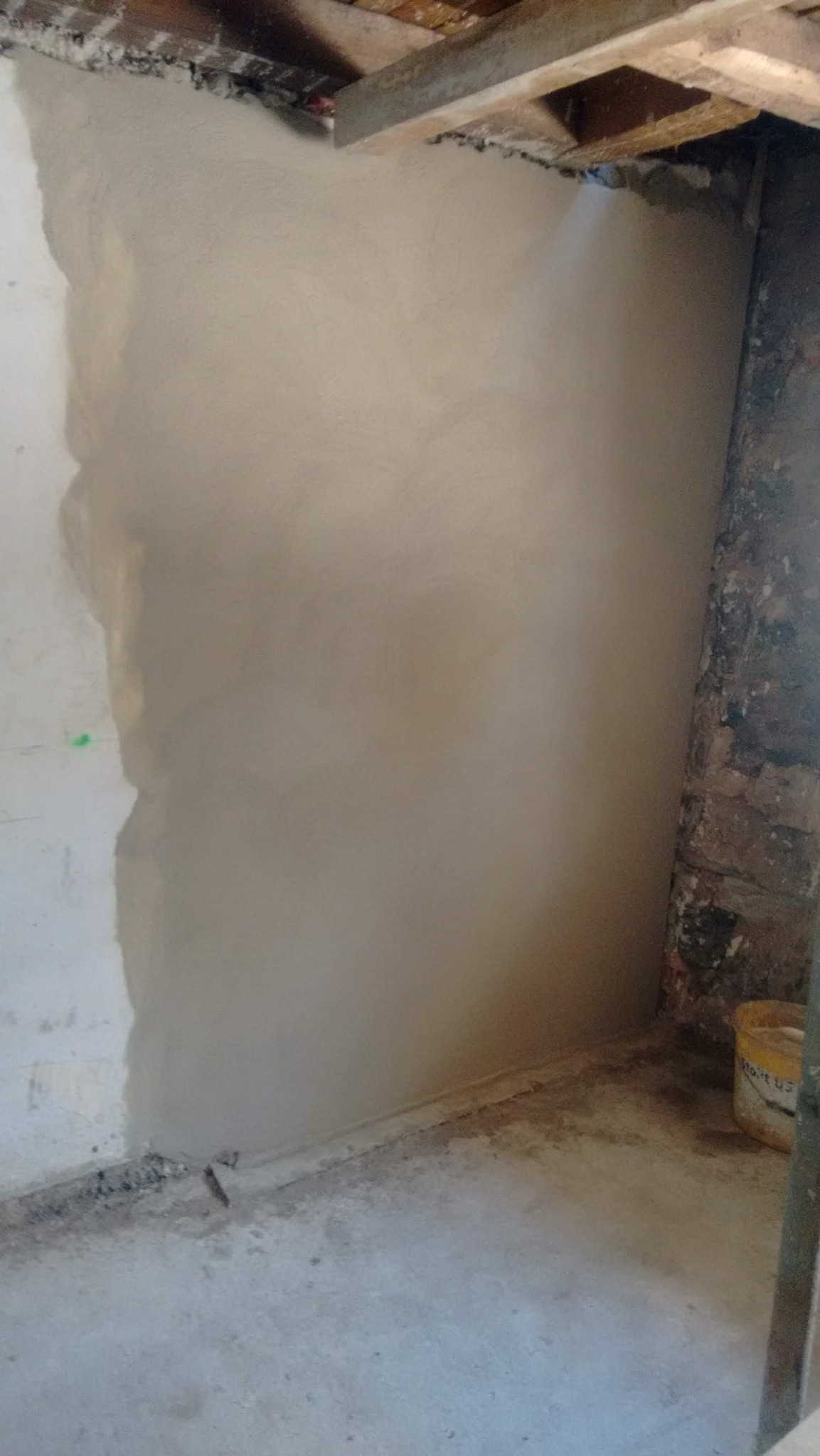 Świeżo nałożona, jasna gładź na ścianie w trakcie remontu. Widoczne surowe drewno stropu i fragment muru. Na podłodze wiadro z zaprawą.