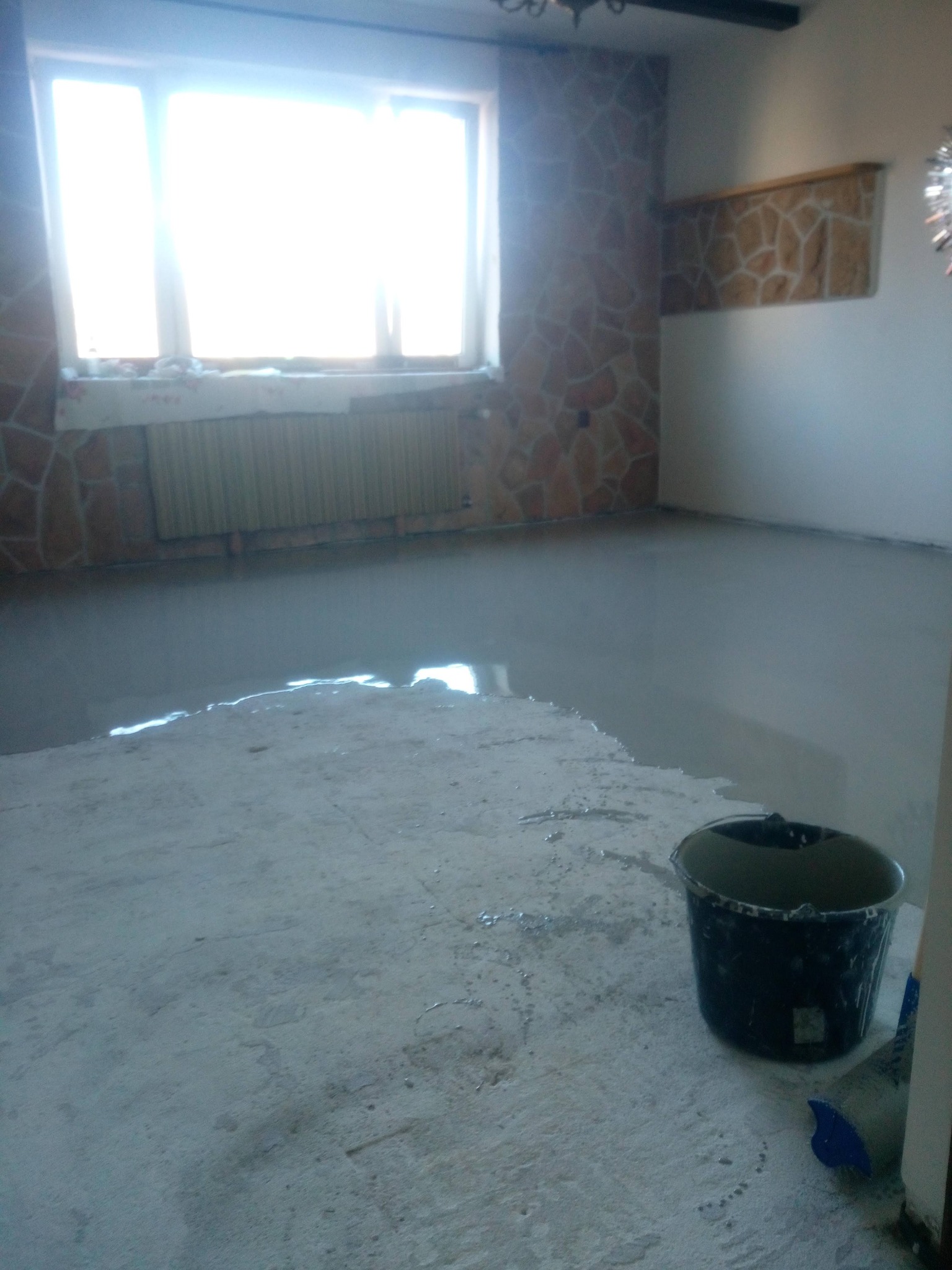 Wylany podkład podłogowy w pokoju z oknem i dekoracyjną okładziną ścienną. Wiadro i narzędzia obok świeżo wylanej posadzki.