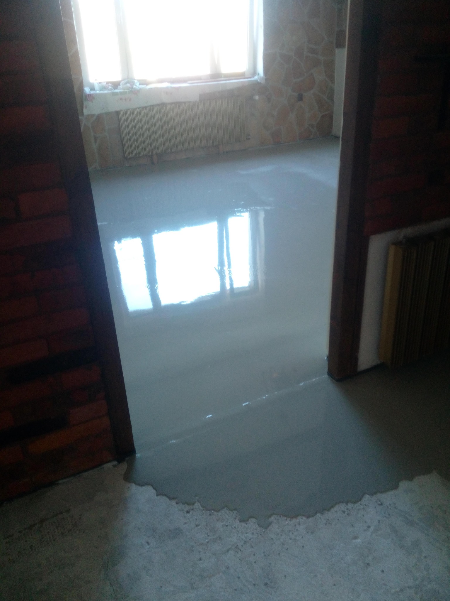 Świeżo wylana, gładka wylewka samopoziomująca na podłodze w remontowanym pokoju, widok przez framugę drzwi. Na ścianie dekoracyjna okładzina kamienna.