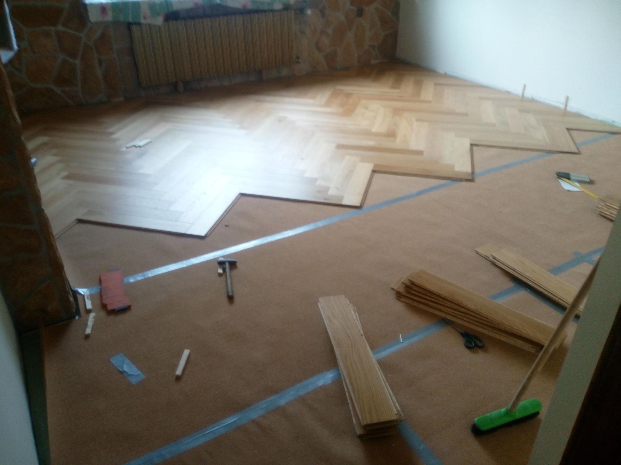 Układanie parkietu w jodełkę na brązowym podkładzie. Widoczne narzędzia: młotek, miarka, nożyczki. Część podłogi już wykończona, reszta w trakcie prac.