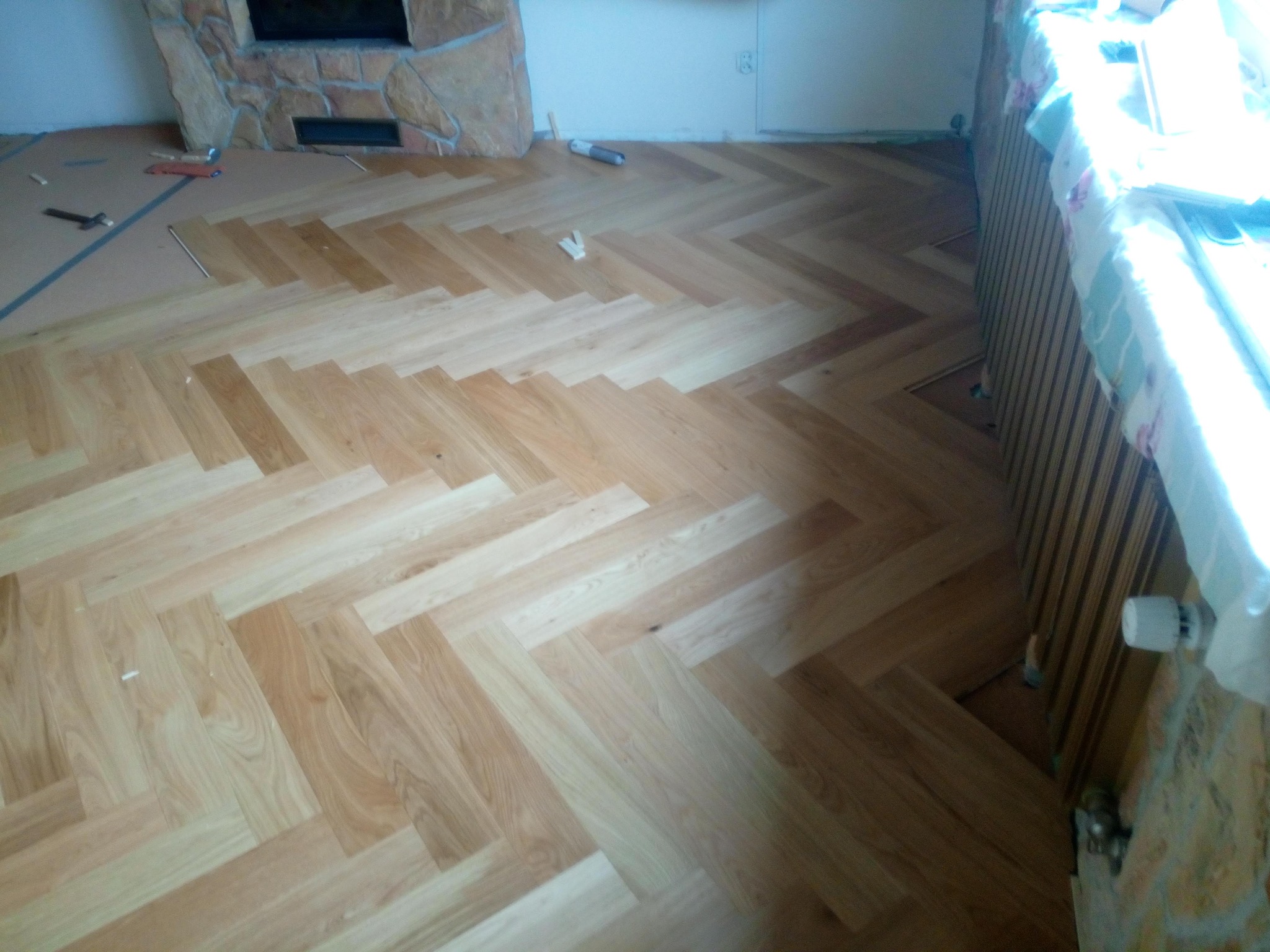 Ułożona podłoga z paneli w jodełkę, widoczne narzędzia i zabezpieczony grzejnik. Częściowo ułożona podłoga w pokoju z kominkiem.
