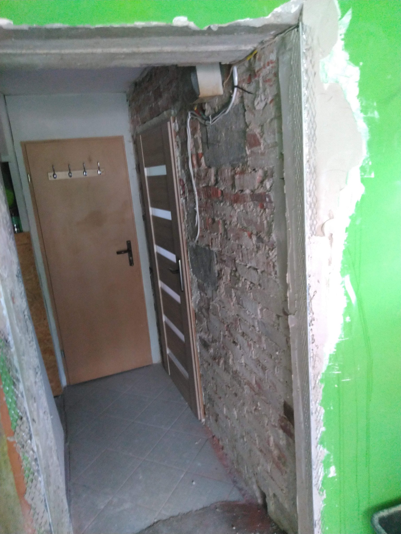 Narożnik ściany z widoczną cegłą i fragmentem nowej instalacji. W tle widoczne drzwi i płytki podłogowe. Prace przygotowawcze przed wykończeniem.