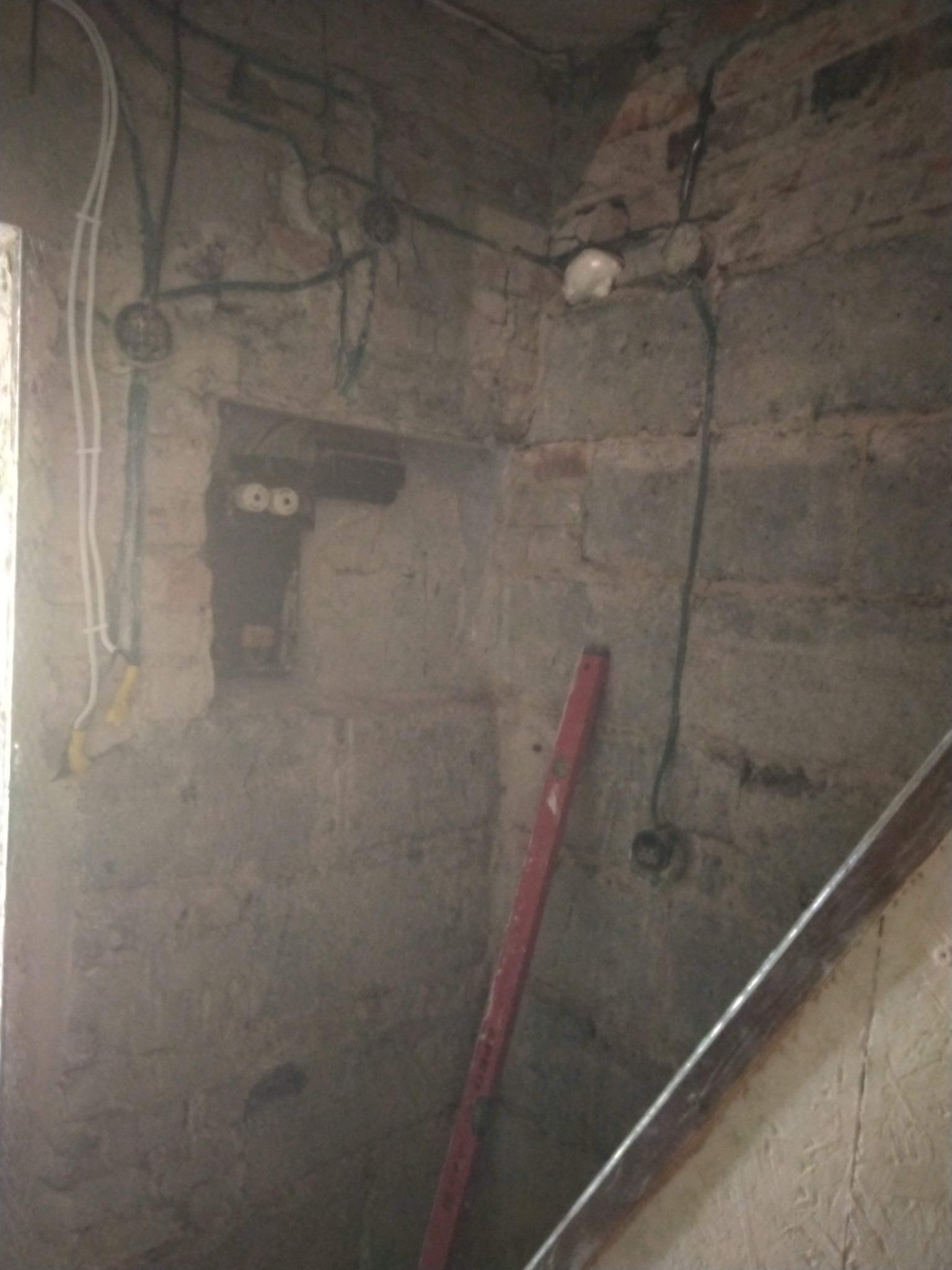 Surowa ściana z cegły z widoczną instalacją elektryczną, puszkami i przewodami. Czerwona poziomica oparta o ścianę. Wnęka w ścianie z elementami instalacji.