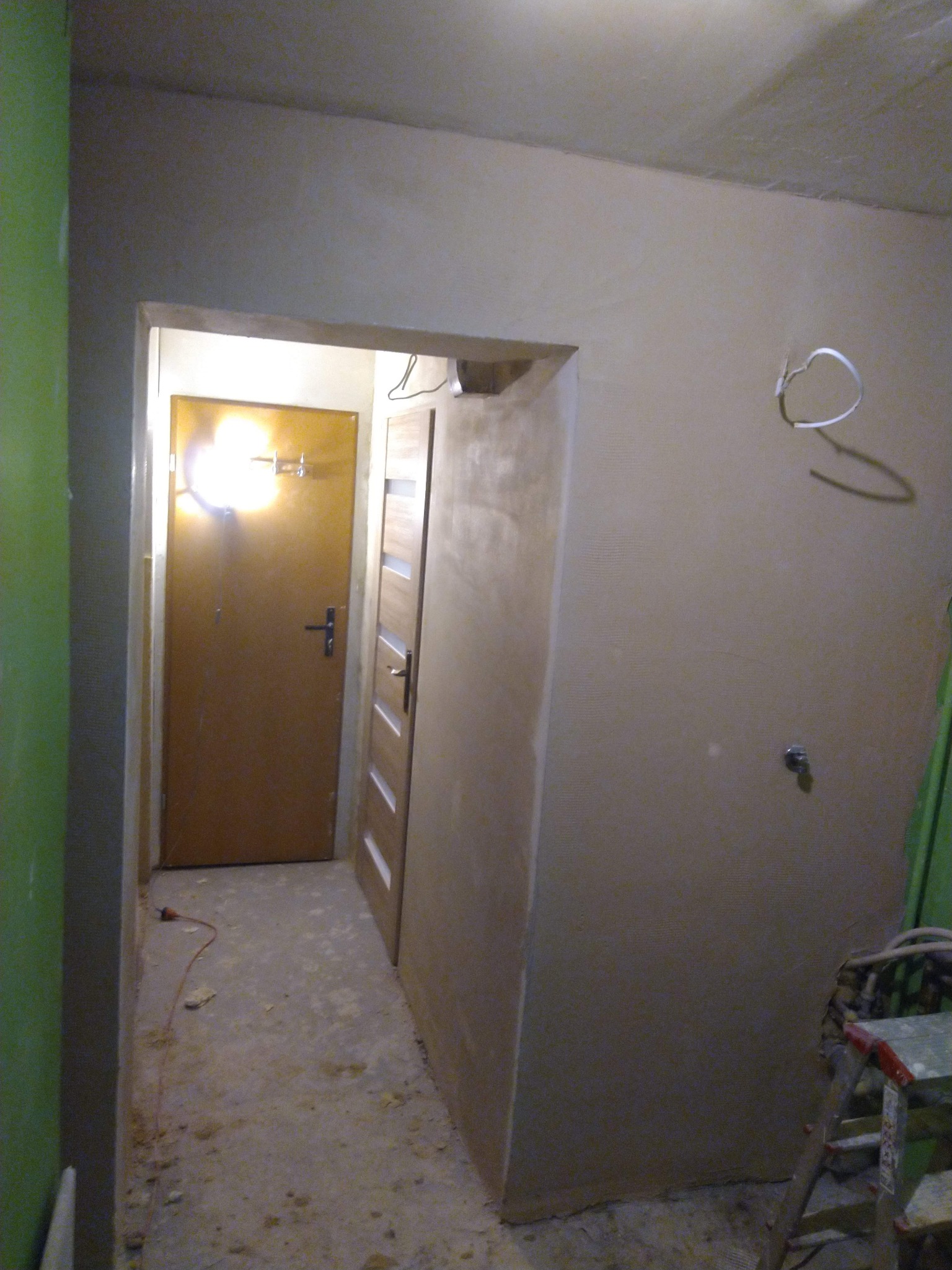 Korytarz w trakcie remontu: ściany po szpachlowaniu, widoczne dwie ościeżnice drzwiowe, kabel na podłodze i drabina malarska. Prace wykończeniowe wewnątrz budynku.