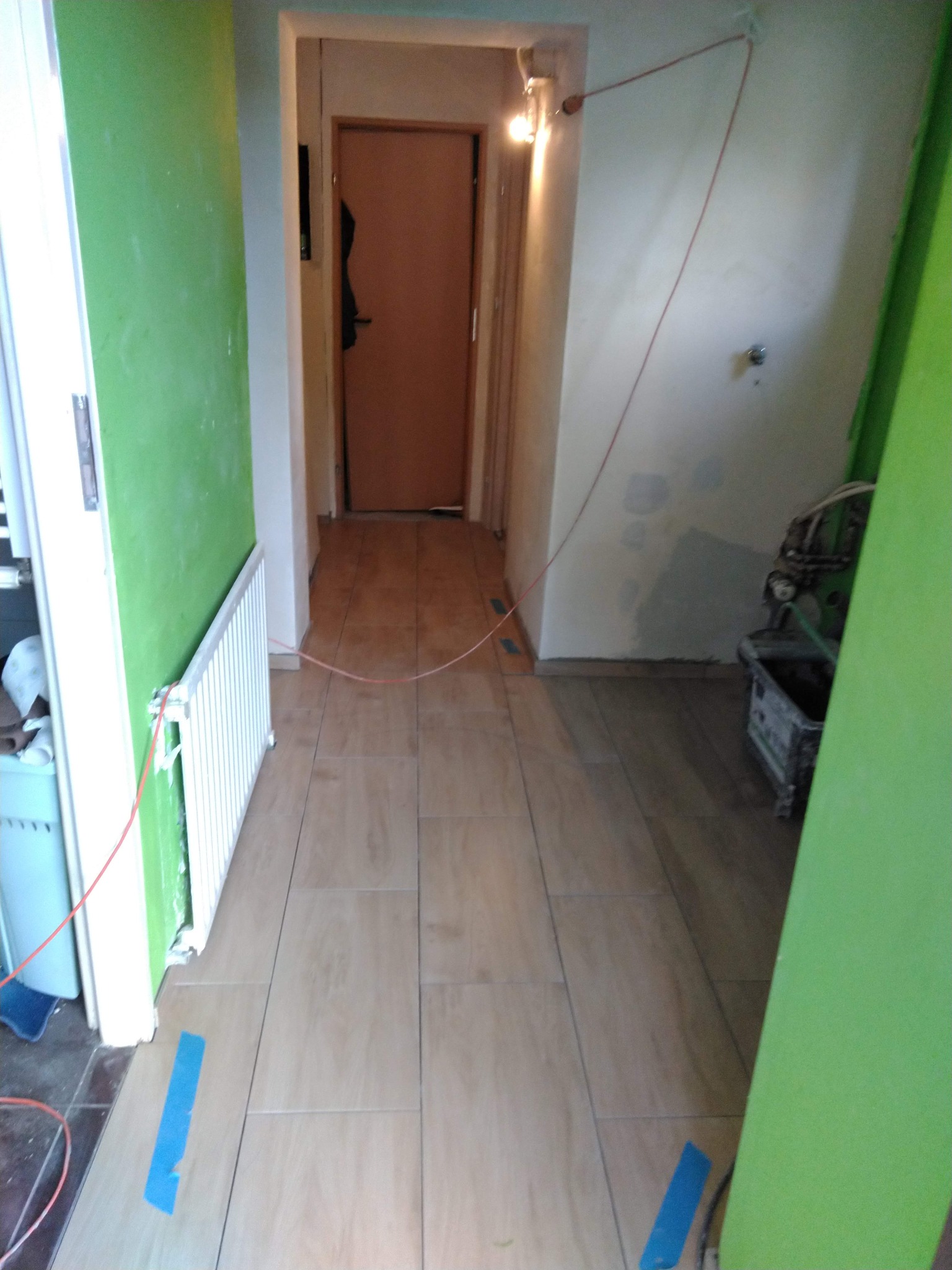 Korytarz w trakcie prac: ułożone płytki podłogowe w odcieniach beżu, ściany częściowo pomalowane na zielono, widoczne narzędzia i taśma malarska.