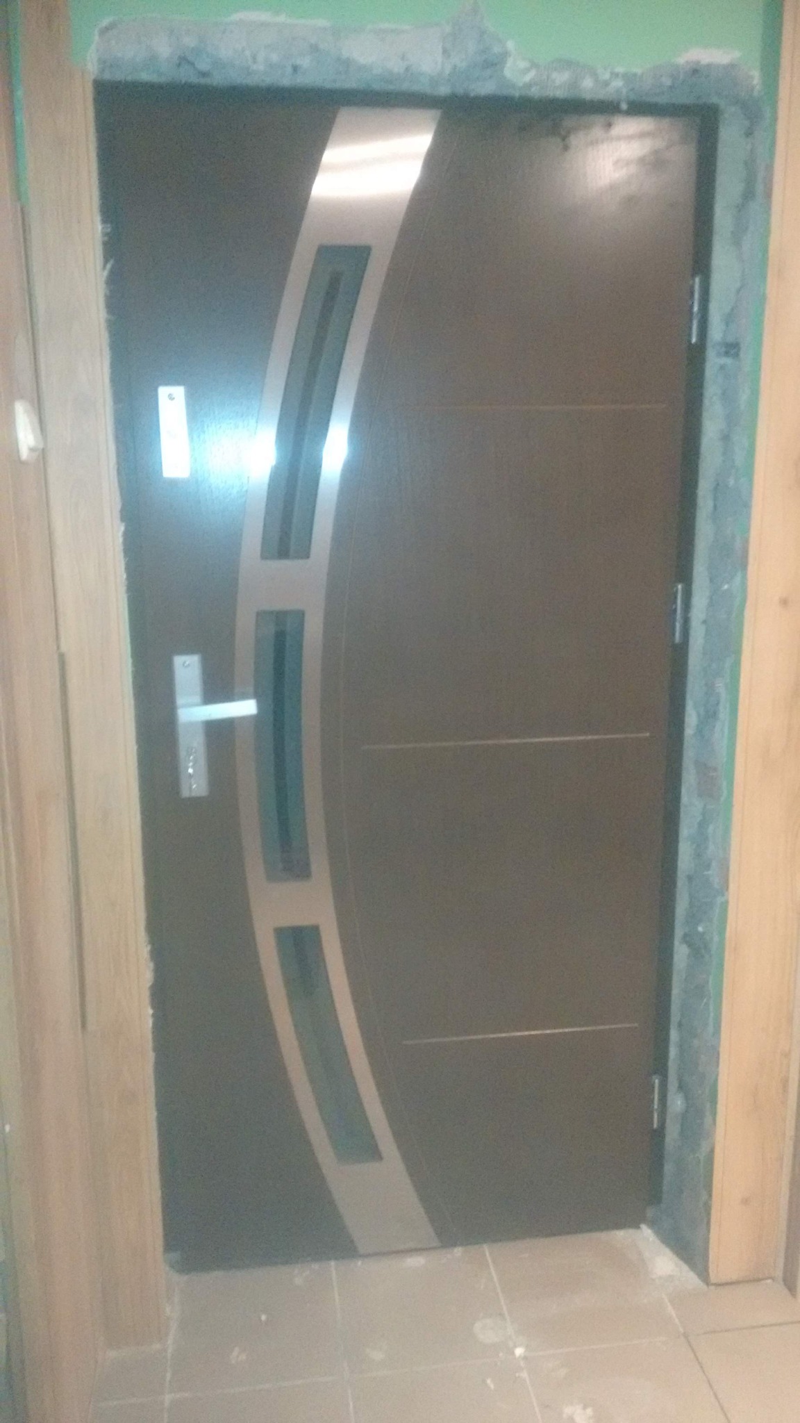 Brązowe drzwi wejściowe z srebrnymi wstawkami i klamką, osadzone w surowym otworze drzwiowym, widoczne płytki podłogowe i fragment ościeżnicy.