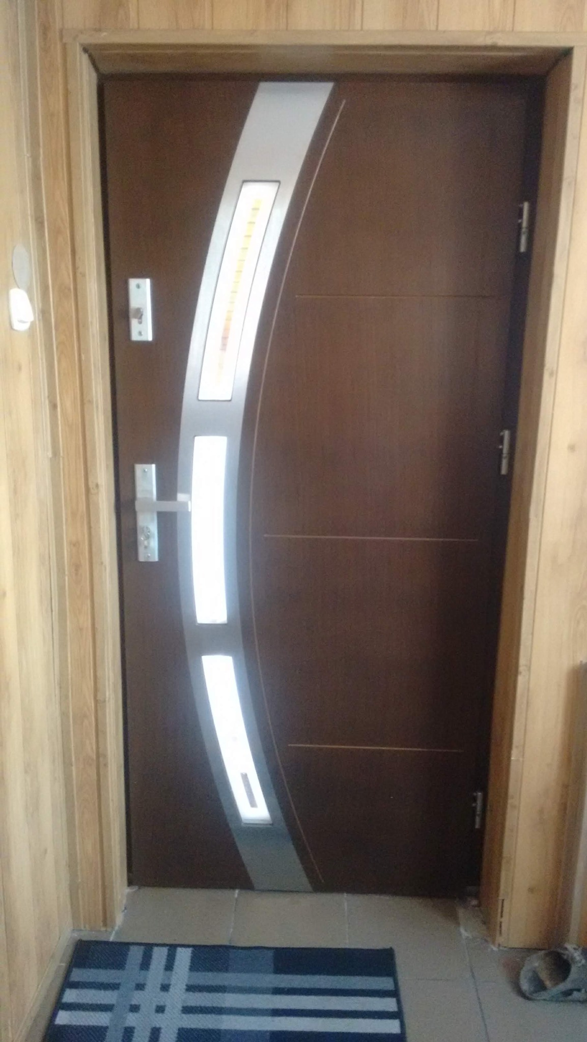 Ciemnobrązowe drzwi wejściowe z dekoracyjnym, srebrnym panelem z podłużnymi przeszkleniami, zamontowane w futrynie. Na podłodze leży wycieraczka.