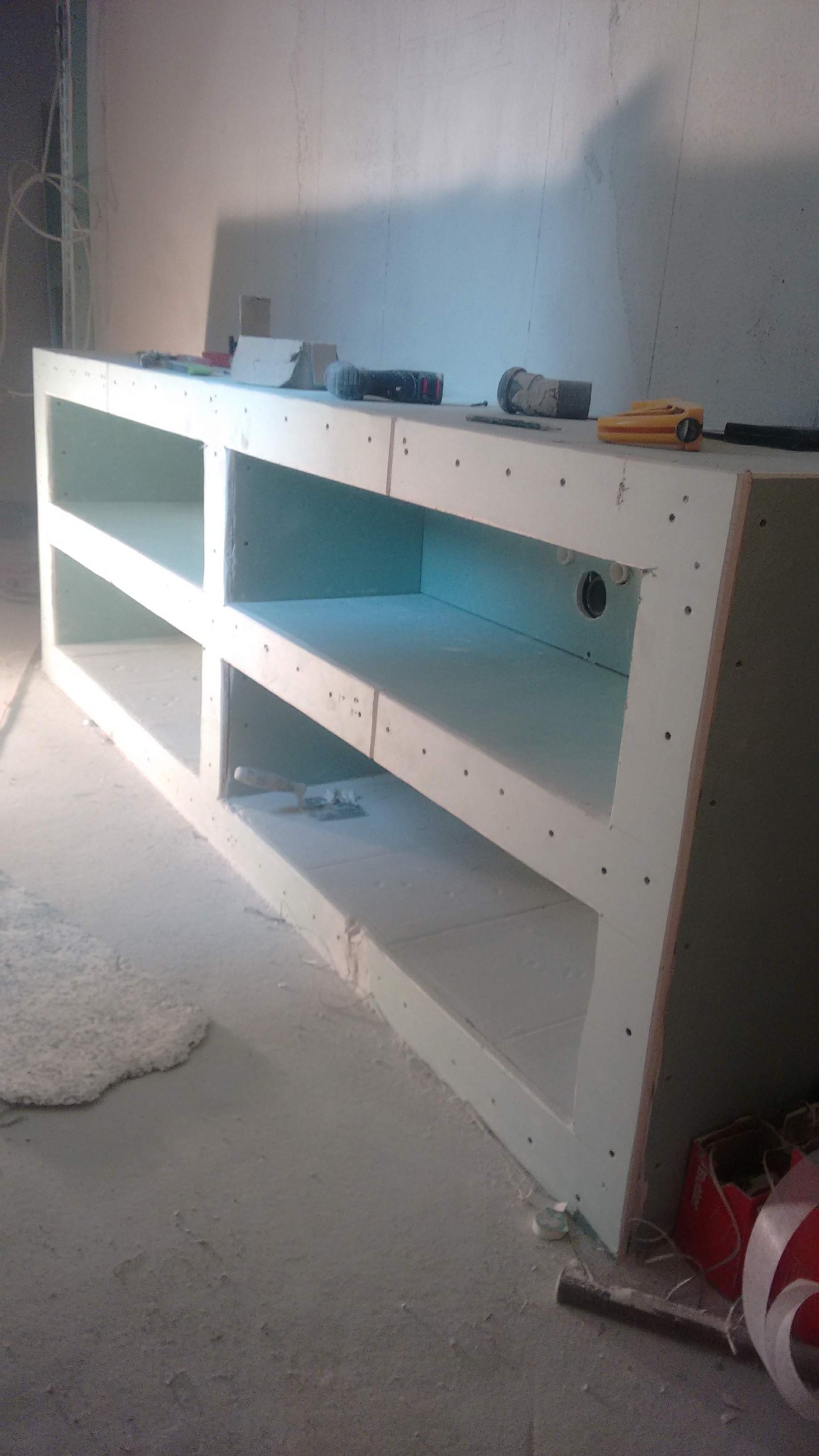 Nierówna powierzchnia ściany za konstrukcją z płyt gipsowo-kartonowych, tworzącą półki. Widoczne narzędzia i akcesoria budowlane. Surowy stan wnętrza.