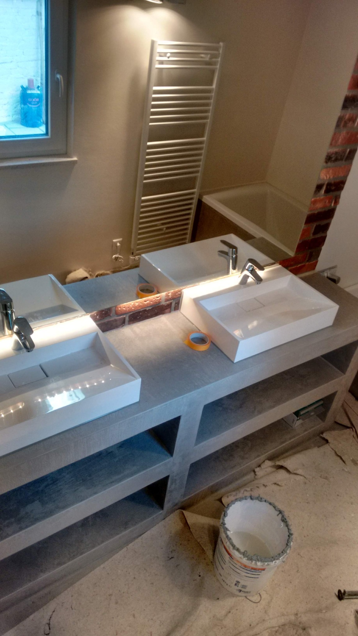 Nowoczesna łazienka z podwójną umywalką na szarym blacie, widoczne taśmy malarskie i wiadro, fragment ściany z cegły w lustrze. Grzejnik drabinkowy w tle.