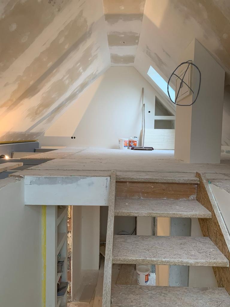 Poddasze w trakcie prac wykończeniowych: surowe ściany, schody z płyty OSB, widoczne narzędzia i materiały budowlane. Okno dachowe w tle.