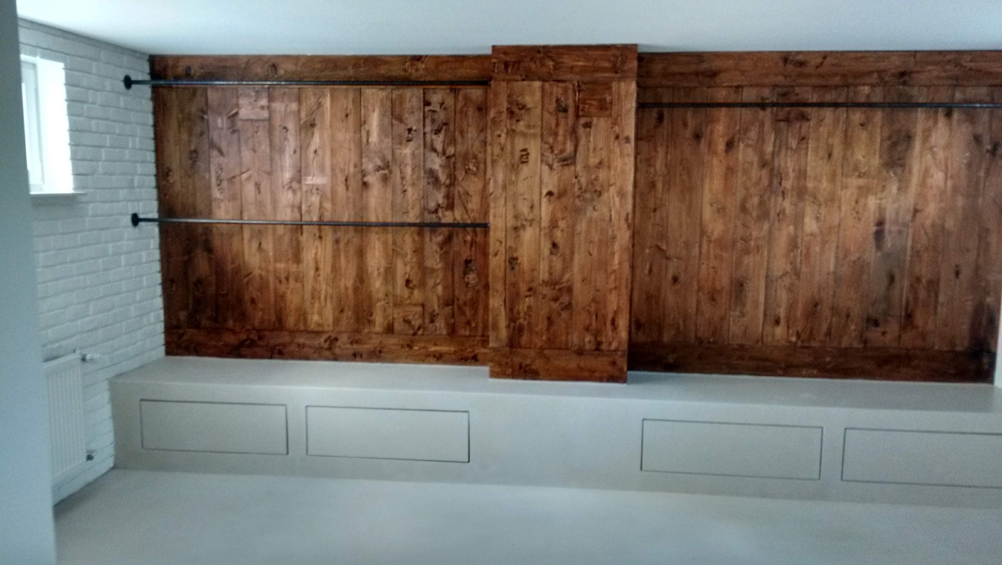 Drewniana ściana z przesuwnymi drzwiami w stylu rustykalnym, uzupełniona metalowymi rurami. Biała cegła i zabudowa z szufladami w jasnym wnętrzu.