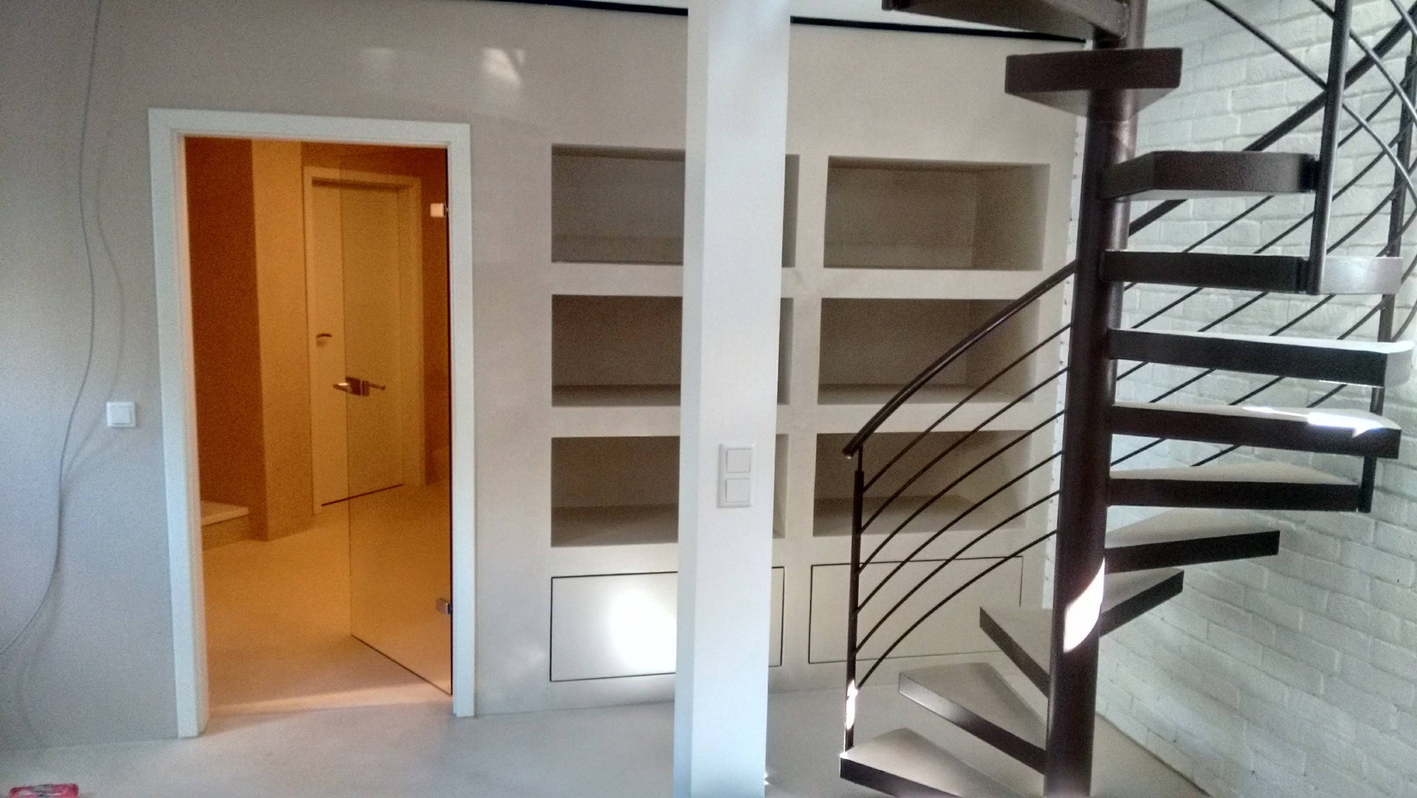 Wykończone wnętrze z spiralnymi schodami, wnękami w ścianie i otwartymi drzwiami. Minimalistyczny design, jasne kolory, ceglana ściana w tle.