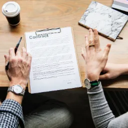 Spotkanie biznesowe przy drewnianym stole z laptopem, filiżanką kawy i notatnikami; dwoje ludzi omawia kontrakt, jeden z długopisem w dłoni, drugi z rękami uniesionymi w geście dyskusji, oboje...