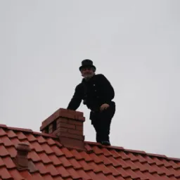 Kominiarz w tradycyjnym stroju (czarny frak i cylinder) stoi na dachu z czerwonej dachówki obok ceglanego komina, z widoczną szczotką kominiarską.